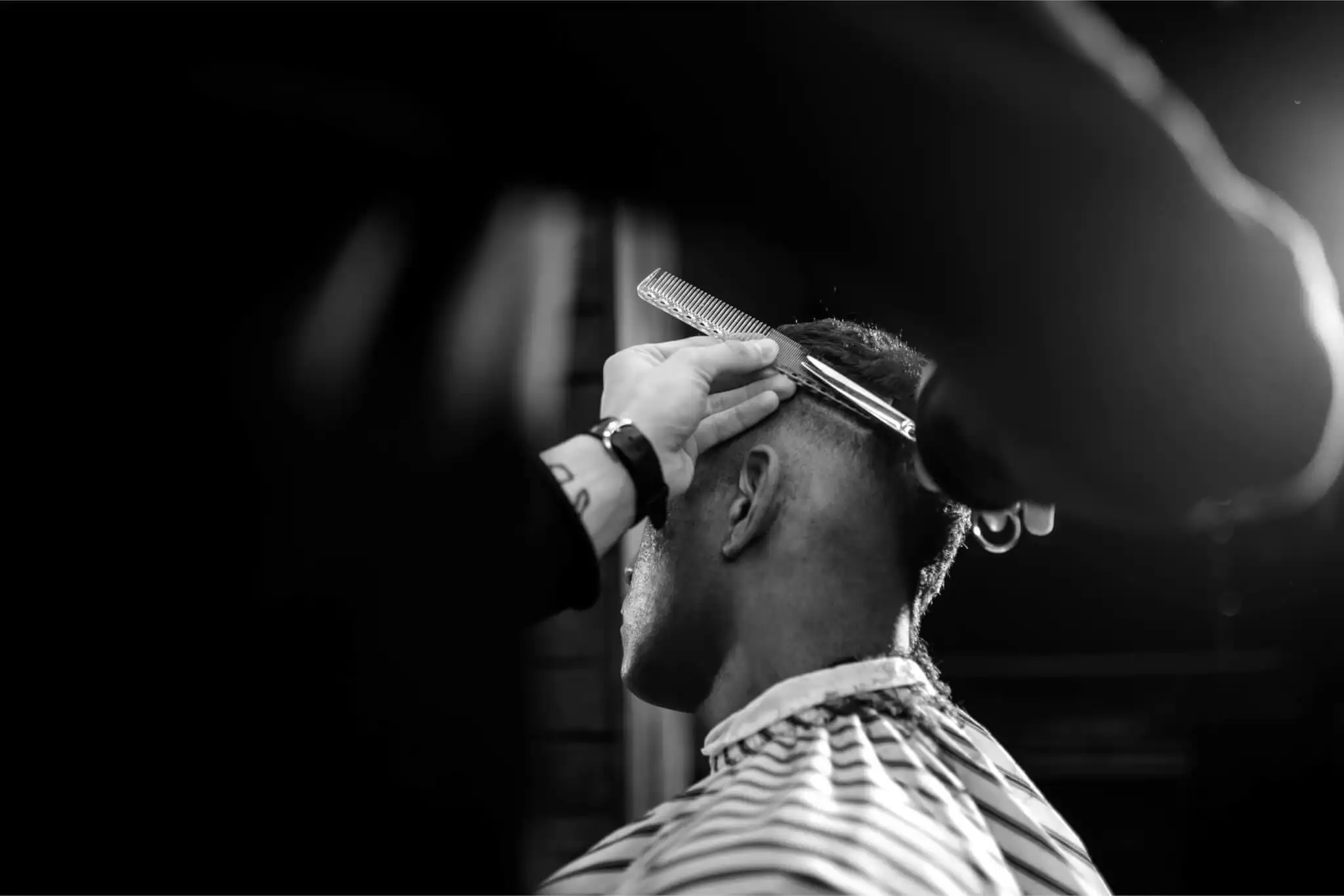 Close-up black and white photo of a barber's hands using a comb and scissors to detail a client's fade.